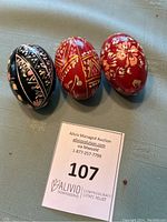 Three hand-painted wooden eggs with European style floral and geometric designs in red and black colors, placed on a surface with a lot number card.