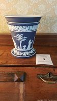 Full view of dark blue Wedgwood vase with white classical relief decoration sitting on wooden surface.