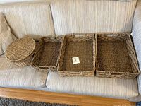 Overview of six woven baskets placed on a beige sofa showing variety of shapes and sizes including rectangular trays, square baskets, and an oval lidded basket.