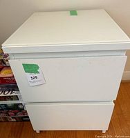 Front view of white bedside table showing two drawers and a green sticker with lot number on the upper drawer.