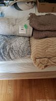 Blanket lot comprising four different blankets shown in a grouped arrangement on a white surface. Colors include beige, brownish taupe, gray with leaf patterns, and tan with chevron puff pattern. Various textures and sizes visible.