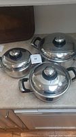 Four polished stainless steel Carrera cooking pots with matching lids and black handles on a kitchen counter.