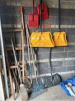 Photo showing rakes, brooms, red scoop shovels, yellow snow shovels, and other garden tools against a wall in a garage or shed.