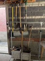 Photo of gardening tools hanging on wall including shovels with wooden and green handles, a garden roller, and wooden handles showing wear.