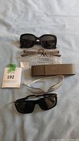 Photo of two sunglasses pairs, clear glasses, brown case, and soft pouch on a fabric surface.