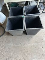 Four black square plastic planter pots stacked and placed together on the ground, visible from a top angle showing the hollow interior and overall structure.