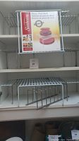 Metal white wire shelf stacking organizers displayed on shelves with price tags. New Sunbeam branded plate shelf organizer boxes show reversible stacking use.