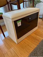 White wooden shoe storage bench with white upholstered seat and black glass or acrylic front panel. Shows bench with front panel closed.