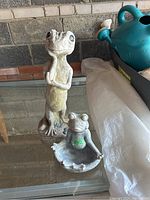 Two stone statues on glass surface: a seated frog on a shell base and a standing gecko.