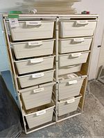 Front view of plastic storage bin unit showing all 10 cream-colored drawers housed in white metal frame, placed on concrete floor with some items on top of the unit.