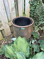 Photo shows a large cylindrical metal planter outdoors, decorated with embossed sunburst patterns on its side, some rust and patina visible. It is surrounded by plants and near a wooden fence.
