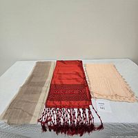 Three scarves laid flat on a white surface showing the rusty-red embroidered scarf with beadwork and tassels in the center, a tan scarf on the left and a pale pink/peach scarf on the right.