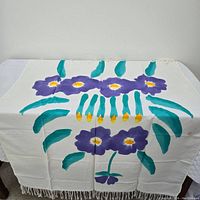 Full view of the rectangular table cloth laid flat showing the bright floral pattern with purple flowers, yellow accents, and green leaves on white fabric with fringe edges.