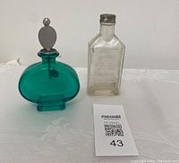 Two decorative glass bottles side by side on a white lace tablecloth with a Maxsold tag