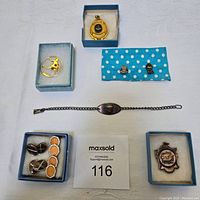 Top-down view of jewellery including gold-tone bracelet with central pendant, brooches, pairs of cufflinks, and shield emblem.