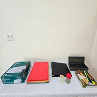Wide shot showing all items on table including envelopes, file folders, black binder, jar of push pins, roll of labels, and metal storage box with key