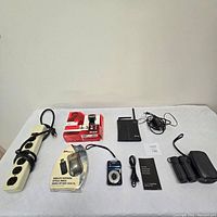 Seven technology and optical items including power extension, wireless booster, mouse, camera, and binoculars on white surface.