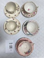 Five vintage china teacups and saucers arranged on a white quilted surface showing various floral patterns including pink roses and gold rims.