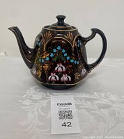 Front and side view of vintage brown ceramic teapot with hand painted floral and gold designs on white tablecloth, showing spout and handle, labeled lot 42.