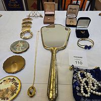 Full set of vintage vanity accessories and jewelry arranged on white cloth, includes mirror, compacts, rings, bracelet, necklaces, and hair combs.