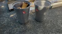 Two aluminum buckets standing upright on a rough surface, one with small items inside