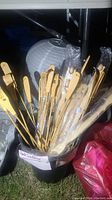 Plastic bucket holding many wooden backscratchers, some wrapped in plastic, some unwrapped, different handle styles visible, placed outdoors.