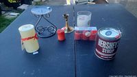 Photo of all candles and candle holders on a black outdoor table, showing Hershey's Syrup candle and multiple other candles and holders.