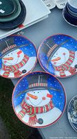 Three ceramic Christmas plates with a snowman wearing a top hat and striped scarf on a blue snowy background, arranged in a triangle shape.