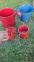 Four ceramic pots arranged on grass: two large, one red and one blue, and two smaller red pots, one held by hand.