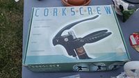 Photo showing a black lever-style corkscrew wine opener in its original box with vintage graphic design and text indicating use for opening wine.