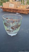 Photo of a clear glass cup decorated with black fish skeleton designs and aquatic plants, with a gold rim on the lip.