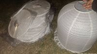 Photo of multiple white paper lanterns in plastic wrapping lying on the ground, showing wireframe and circular shape with openings.