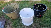 Three ceramic plant pots displayed outdoors on grass: beige with teardrop cutouts, white with drainage holes and saucer, black with leaf cutouts.