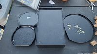 Four black plastic trays arranged on a surface, including one rectangular and three round trays of varying sizes.