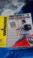Box showing Wagner Power Painting System 695E with image of man wearing backpack sprayer unit painting exterior wall, highlighting electric paint spraying system.