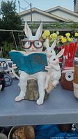 Rabbit ornament seated on a stump wearing glasses, holding a blue book with a smaller rabbit touching the book.