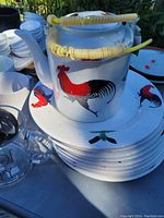 Teapot with yellow woven handle sits atop a stack of ceramic plates decorated with red and black roosters.
