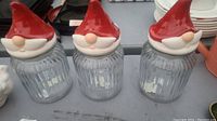 Three clear ribbed glass jars each with a ceramic Santa Claus head lid featuring a red hat, beige nose, and white beard.