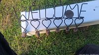Photo of black metal wire welcome sign with seven small rusty bells hanging from heart-shaped hooks, placed on grass.