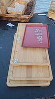 Photo shows a stack of three light-colored wooden trays with a smaller red tray on top, all on a black surface with some wicker baskets and a bamboo cage item in the background.