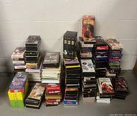 Photo showing multiple stacks of VHS tapes arranged on floor against a white wall. Various movie titles and genres visible on tape sleeves.