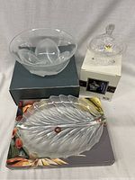Three boxed glassware items consisting of a large clear bowl on a black box, frosted leaf-shaped dish with a bronze accent on a floral box, and ribbed covered candy dish on its white original box.