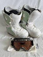Pair of Burton women's snowboard boots white and light green next to Oakley goggles with amber lens and white soft bag