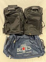 Three duffel bags: two large black Samsonite duffel bags with telescopic handles and wheels, and one blue University of Toronto Varsity Blues sports duffel bag.