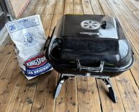 Viewing the closed Grill Smith black tabletop charcoal grill with vent on lid, handle, and foldable legs on wooden floor next to a bag of Kingsford charcoal briquets.