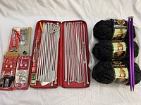 Full layout of vintage Areo knitting needles in red zip case, newer circular needles in packaging, large purple needles, three balls of black chunky Wool-Ease yarn and various sewing accessories.