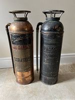 Pair of vintage copper and brass fire extinguishers standing side by side on tile floor, showing full height and body condition.