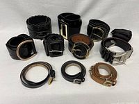 All ten belts laid out, showing variety in buckle design and leather finish: black and brown leathers, metal buckles in silver and gold tones.