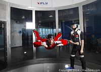 Man flying horizontally in iFly wind tunnel in red flight suit with black helmet and instructor standing nearby in black and red suit inside wind tunnel.