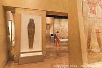 Photo of an exhibit room displaying an ancient Egyptian sarcophagus in a glass case with a visitor walking by.